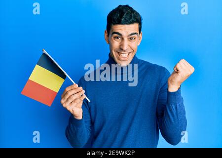 Handsome hispanic man holding belgium flag screaming proud, celebrating victory and success very excited with raised arm Stock Photo