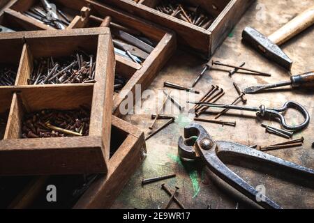 Vintage work tool and toolbox on wooden table. Messy industrial equipment in carpentry workshop. Pliers, screwdriver, hammer, hand drill and many nail Stock Photo