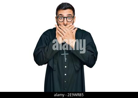 Young hispanic man wearing priest uniform shocked covering mouth with hands for mistake. secret concept. Stock Photo