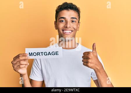 Young handsome african american man holding mortgage word smiling happy and positive, thumb up doing excellent and approval sign Stock Photo