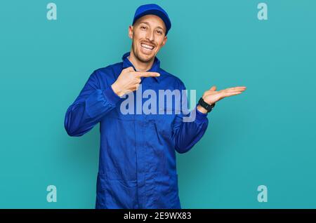 Bald man with beard wearing builder jumpsuit uniform amazed and smiling to the camera while presenting with hand and pointing with finger. Stock Photo