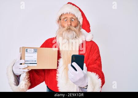 Old senior man with grey hair and long beard wearing santa claus costume holding delivery box and phone making fish face with mouth and squinting eyes Stock Photo