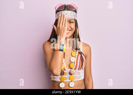 Beautiful hispanic woman wearing bohemian and hippie style covering one eye with hand, confident smile on face and surprise emotion. Stock Photo