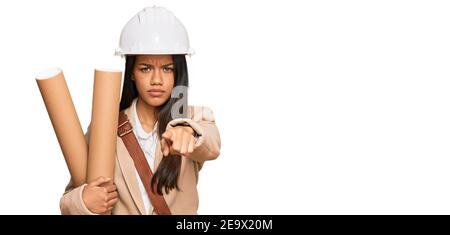 Beautiful hispanic woman holding paper blueprints pointing with finger to the camera and to you, confident gesture looking serious Stock Photo
