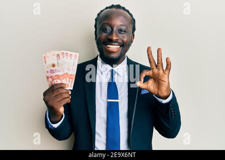 Handsome young black man wearing business suit holding 10 colombian pesos doing ok sign with fingers, smiling friendly gesturing excellent symbol Stock Photo