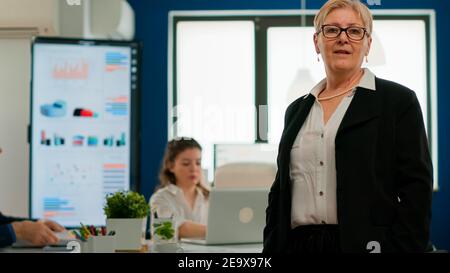 Portrait of successful senior entrepreneur standing in front of meeting desk looking at camera. Manager working in professional startup financial business modern company workplace ready for conference Stock Photo