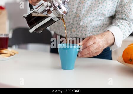Older man spilling coffee in cup after making it using french press for breakfast in kitchen. Elderly person in the morning enjoying fresh brown cafe espresso cup caffeine from vintage mug. Stock Photo