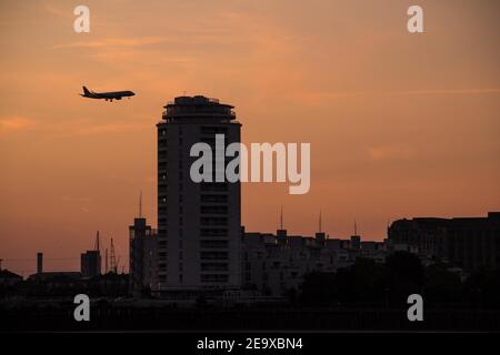 A silhouetted airplane landing at London City Airport, against the orange glow of sunset.