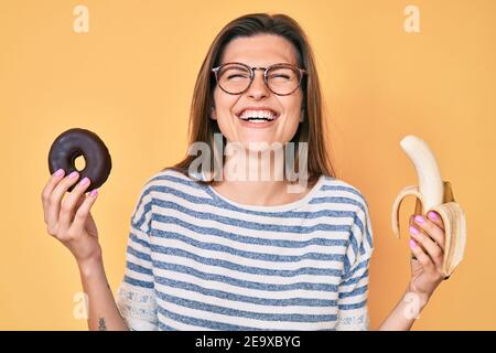 Beautiful caucasian woman holding banana and chocolate doughnut smiling and laughing hard out loud because funny crazy joke. Stock Photo