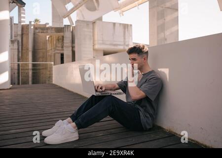 Full body side view of young concentrated male freelancer in trendy outfit working remotely on laptop sitting on terrace on sunny day Stock Photo