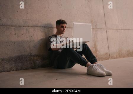 Full length of serious young self employed guy in trendy outfit using laptop while sitting on ground leaning on concrete wall during remote job Stock Photo