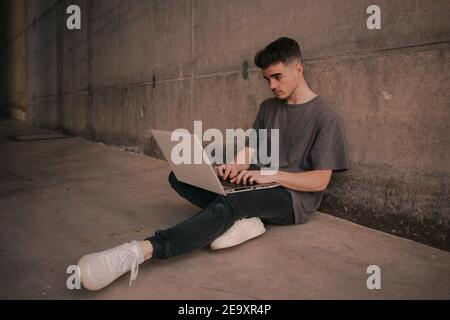 Full length of serious young self employed guy in trendy outfit using laptop while sitting on ground leaning on concrete wall during remote job Stock Photo