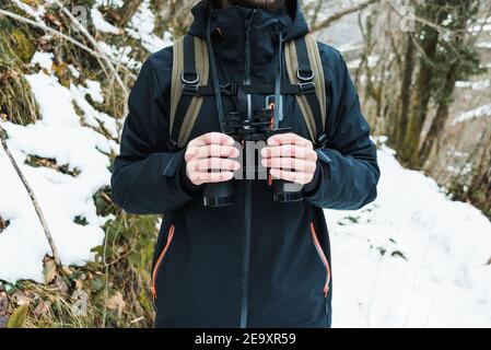 Cropped unrecognizable male hiker in warm clothes standing in snowy winter woods with binoculars Stock Photo