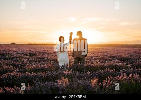 Back view of faceless newlywed couple wearing classy wedding gowns dancing and holding hands gently on spacious blooming lavender field at picturesque Stock Photo