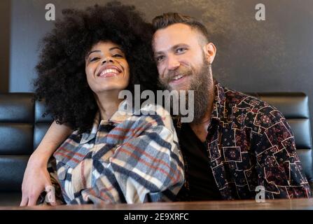 Positive multiracial couple sitting at table in cafe and hugging while smiling and looking forward Stock Photo
