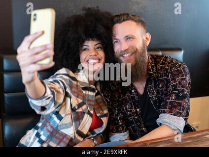 Delighted multiracial couple sitting at table in cafe and taking selfie on smartphone Stock Photo