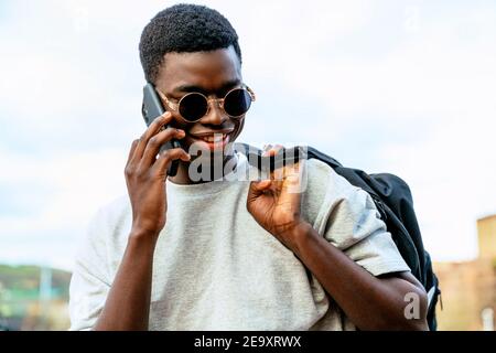 Young African American man with backpack and fashionable sunglasses talking on a cell phone under a cloudy sky. Stock Photo