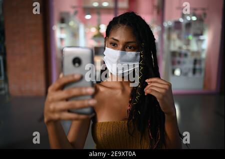 Portrait of attractive young afro latin Woman wearing a facemask takes selfie with smartphone in a commercial mall, Colombia Stock Photo