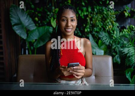 Portrait of attractive young afro latina with dreadlocks in a crochet red top posing with smartphone in restaurant table, Colombia Stock Photo
