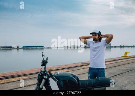 Thoughtful young ethnic unshaven male in casual outfit and cap standing near modern motorcycle with hands behind head and looking away near port Stock Photo