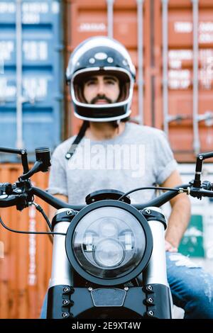 Handsome young ethnic bearded guy in t shirt and helmet sitting on modern motorbike near heap of shipping containers in port and looking away Stock Photo