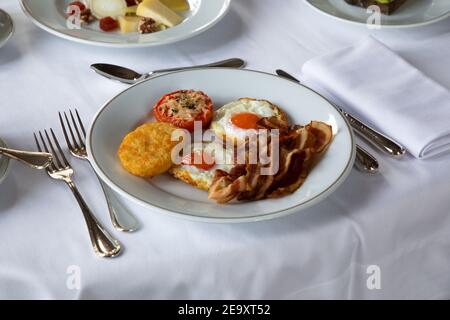 High angle of appetizing fried eggs with bacon slices served on plate with stuffed tomato and crispy mozzarella and placed on table with cutler and cu Stock Photo