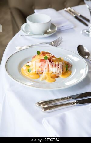 From above of appetizing Eggs Benedict dish with bacon topped with hollandaise sauce served in white plate on table with cup of coffee and silverware Stock Photo