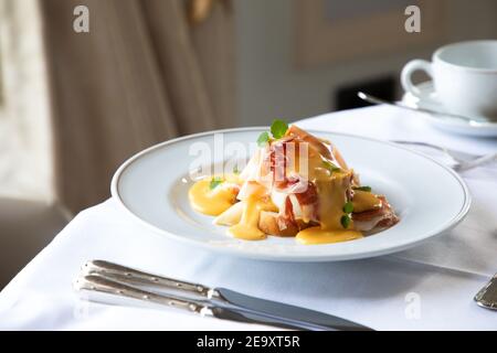From above of appetizing Eggs Benedict dish with bacon topped with hollandaise sauce served in white plate on table with cup of coffee and silverware Stock Photo