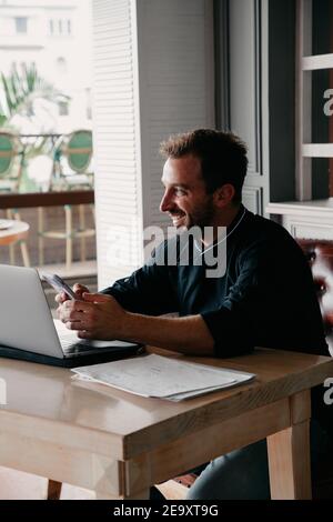 Smiling bearded male chef surfing cellphone while working with netbook at table in restaurant Stock Photo