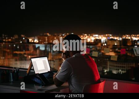 Back view of thoughtful young male freelancer in casual outfit sitting on modern building rooftop and working remotely on laptop in evening Stock Photo