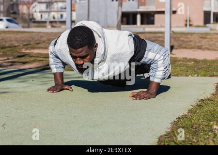 Young fit bearded ethnic male athlete doing push ups looking forward during training on sports ground in town Stock Photo