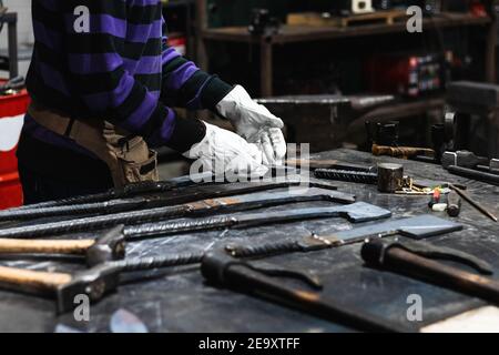 Crop anonymous blacksmith in gloves working in grungy forge with various metal tools Stock Photo