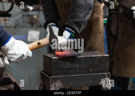 Crop unrecognizable male masters with hammer hitting hot metal detail placed on anvil in forge Stock Photo