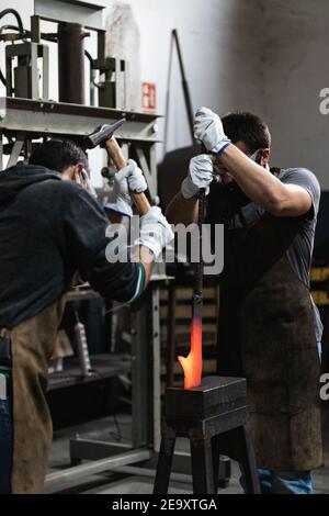 Blacksmiths in aprons forging hot iron with hammer while making metalwork in smithy Stock Photo