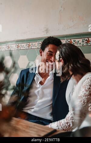Cheerful young multiracial bride and groom sitting at table with flower bouquet and flaming candles while looking at each other in restaurant Stock Photo