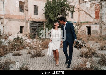 Young glad multiracial groom and bride looking at each other while strolling on walkway against aged construction on wedding day Stock Photo