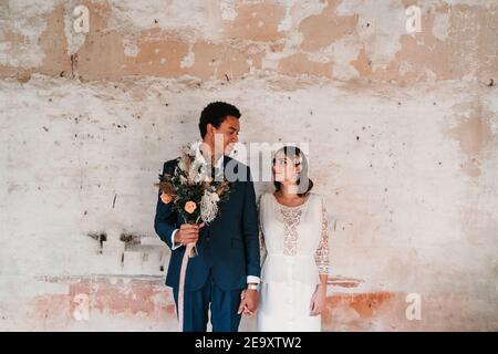 Young smiling multiracial groom with bridal bouquet holding beloved by hand while looking at each other near old wall Stock Photo
