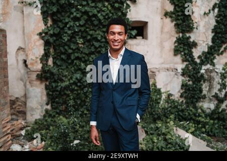 Happy ethnic male in elegant suit standing with hand in pocket and looking at camera against weathered building wall Stock Photo