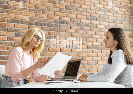 Female middle-aged recruiting officer is interviewing young woman in the office, job seeker. Mature female mentor and a student, trainee indoor Stock Photo