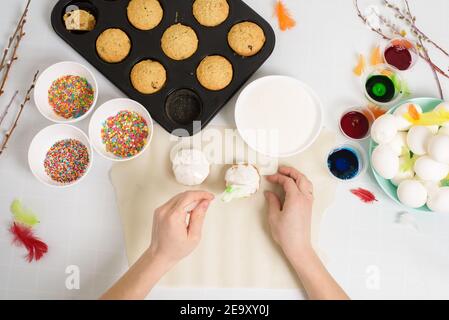 the process of decorating mini cupcakes Easter cakes with white icing and sweet candies, top view, willow branches and eggs for coloring. Stock Photo
