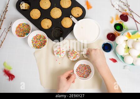 the process of decorating mini cupcakes Easter cakes with white icing and sweet candies, top view, willow branches and eggs for coloring. Stock Photo