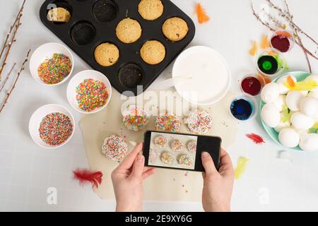 woman takes photos on her phone of mini cupcakes cakes for Easter with white icing and sweet candies, top view, willow branches and eggs for coloring Stock Photo