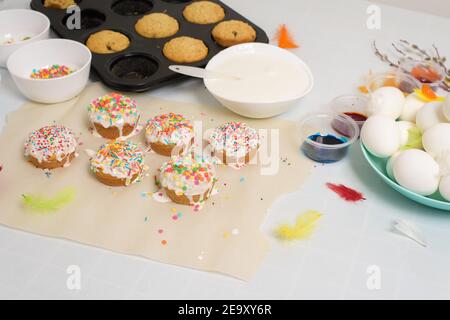 the process of decorating mini cupcakes Easter cakes with white icing and sweet candies, top view, willow branches and eggs for coloring. Stock Photo