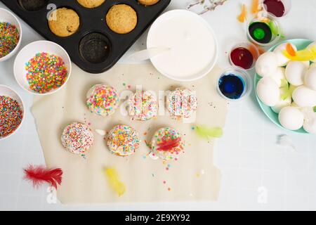 the process of decorating mini cupcakes Easter cakes with white icing and sweet candies, top view, willow branches and eggs for coloring. Stock Photo