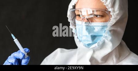 Doctor in personal protective equipment (PPE) holds syringe for COVID-19 vaccine injection. Portrait of nurse wearing face mask and goggles. Concept o Stock Photo