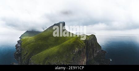 Aerial panorama of a small white lighthouse located on the edge of a huge cliff and the island of Kalsoy. Kalsoy is an isolated small island in the Stock Photo
