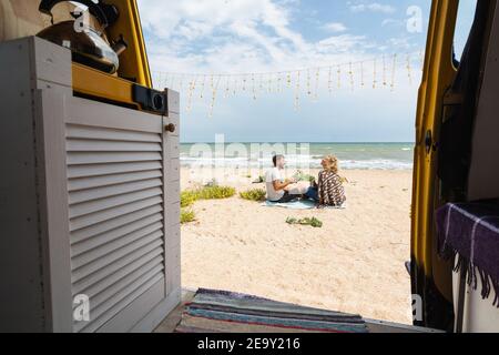 Young Eastern European couple traveling by yellow camper van along the seaside and chilling out at the beach. Self-built off-grid motorhome camping i Stock Photo