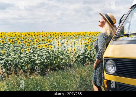 Young blonde woman travelling by campervan. Overlooking sunflower field on sunny summer day. Stock Photo