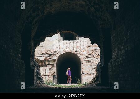 Woman with backpack stands among the ruins of ancient building. Brave solo raveller exploring abandoned ruins. Stock Photo