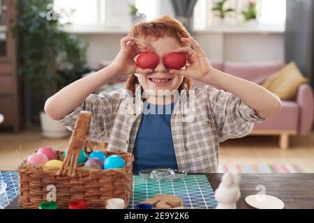 Portrait of boy holding Easter eggs in front of his eyes and smiling at camera while sitting at the table with basket of eggs Stock Photo